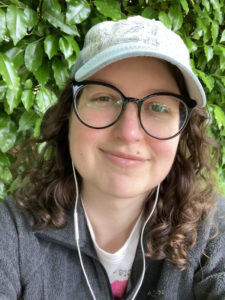 Lizzie has curly brown hair and glasses. She is wearing a cap and standing in front of a green background.