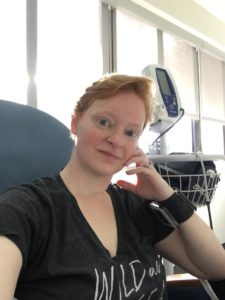 Kelly sits in a medical chair with a vitals machine behind her. She is wearing a black shirt and has a neutral expression.