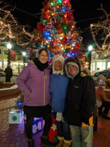 Annemarie stands in front if an outdoor Christmas tree. Her daughter is on her right and her brother is on her left. 