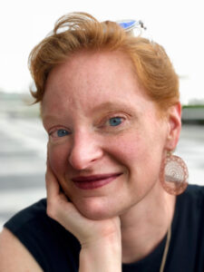 Kelly's author photo. Kelly has reddish-blond hair. She is wearing her hair in a pony tail and has dangly earrings. She is wearing a black shirt. The background is blurred. 