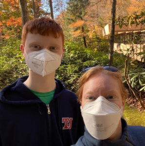 There is a picture of Kelly standing next to her teenage son. Her son is ten inches taller than her. They are both wearing masks and standing in front of some plants. 