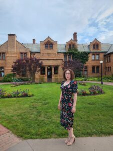 The author is standing outside. She had dark, curly hair and is wearing a black and red dress. There is a large brown house behind her. 