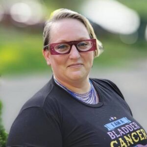 The blogger is a blond woman with red glasses. Her facial expression is neutral. She is wearing a shirt that says "I am a bladder cancer survivor."