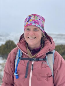 A photo of the blogger. A woman with blond hair in braids wearing a coat, hat, and hiking backpack. She is smiling.