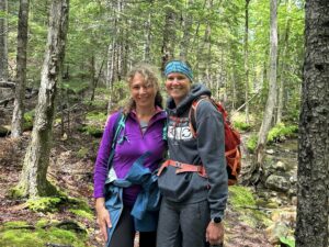 A photo of two women in the woods. They are both wearing sweatshirts and hiking backpacks. They are smiling.