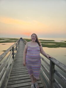A white woman with light brown hair stands on a dock. She is wearing a striped dress. 