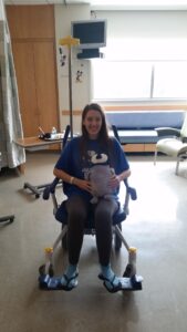 A teenager with brown hair sits in a wheelchair. She is holding a stuffed penguin. 