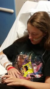 Photo: A girl with brown hair sitting in a hospital bed with and NG tube and IV. 