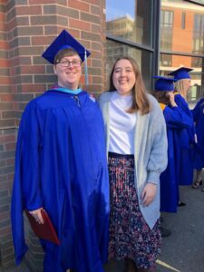 Emily stands next to her fiancé, who is searing a blue cap and gown. Emily is searing a white shirt and a colorful skirt with flowers.