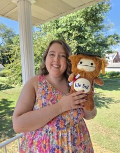 The author, Emily, is wearing a brightly colored dress and holding a stuffed animal with a graduation hat. The stuffed animal says, "We out" on its belly. 